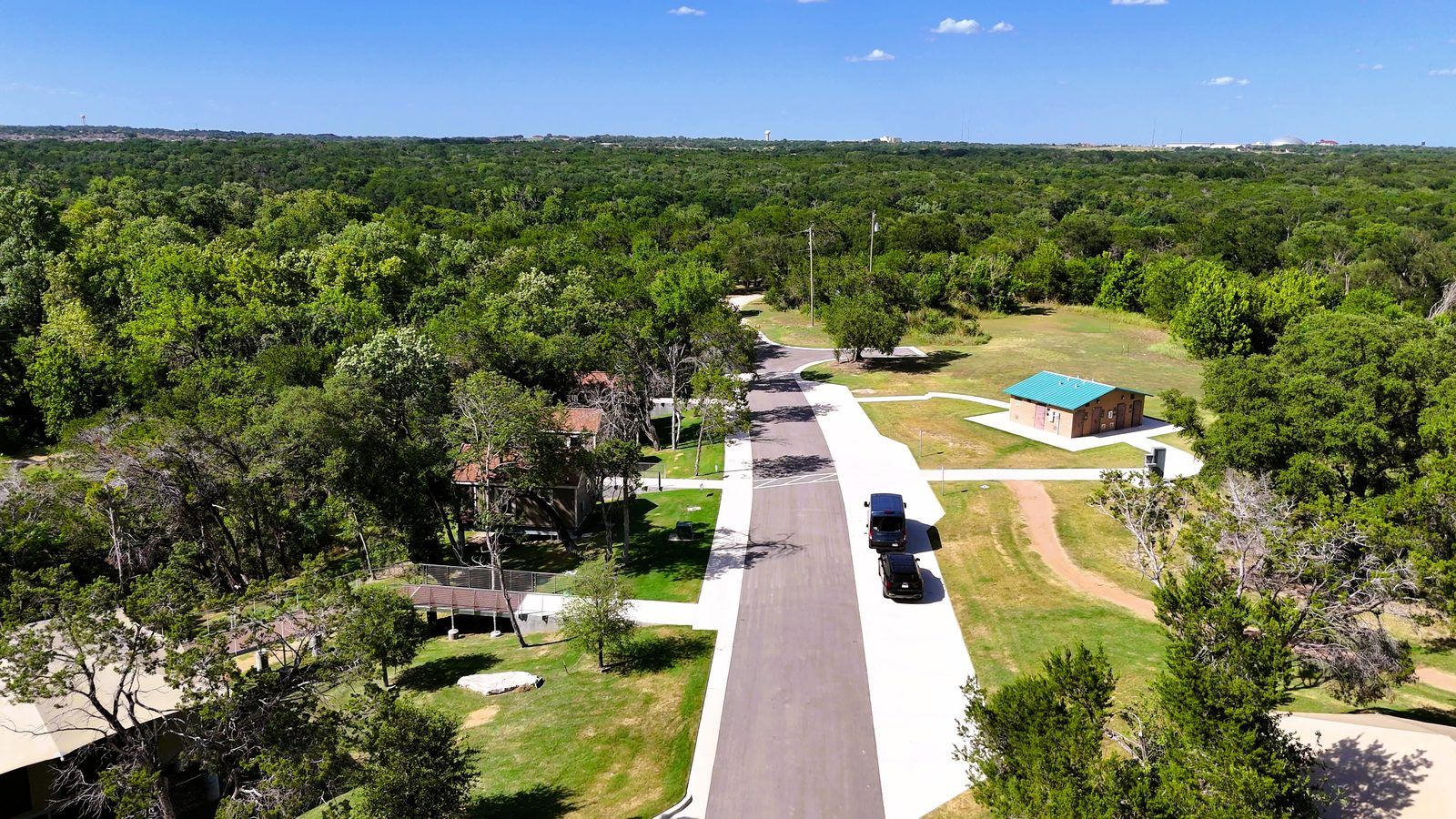 Aerial view of a paved camp road, two parked vans, and a restroom building among trees.